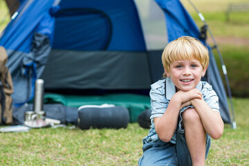 Blond-haired boy kneeling on grassy field at campsite handling thermos beside dome tent, copy space