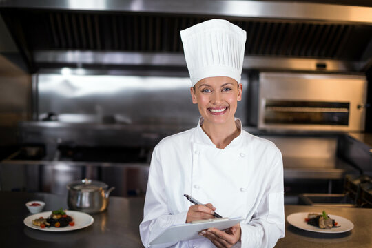 Adult female chef wearing uniform and hat at counter in kitchen holding clipboard inspecting dishes