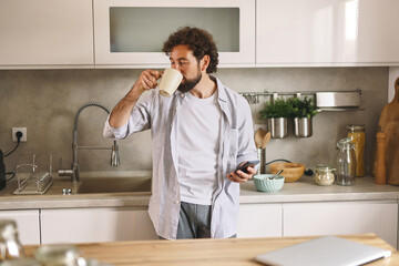 A man stands in a contemporary kitchen, sipping coffee from a mug and looking at his phone. He is casually dressed and surrounded by kitchenware, creating a relaxed atmosphere.