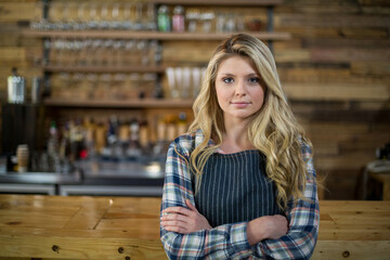 Female bartender wearing plaid shirt and striped apron arranging liquor bottles on wooden counter