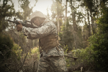 Soldier wearing tactical vest and helmet, moving through dense woodland, scanning ahead with rifle