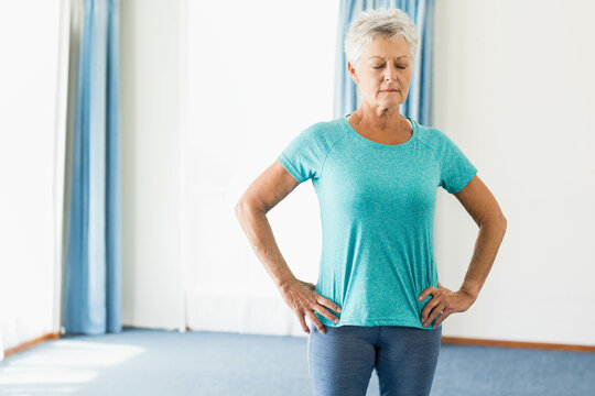 Fototapeta Senior female in gym clothes doing breathing exercise at home by windows with blue curtains