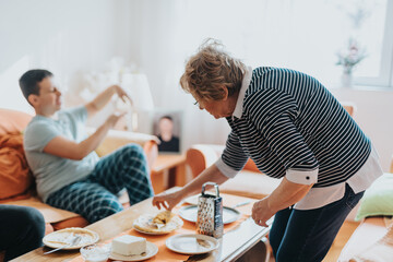 A senior woman arranges food at a living room table while a younger man relaxes in the background, creating a warm and homely atmosphere.