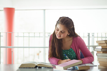 Female teenager writing in book at library table with textbooks and pen near red column railing