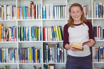 Female child reading open book while standing by library bookshelf with hardcover books, copy space