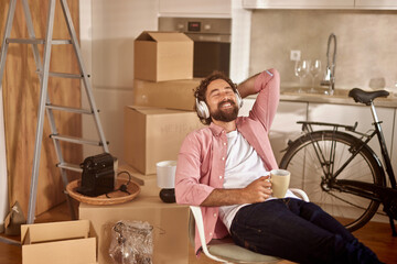 A man with curly hair sits in a chair in his new home, smiling while wearing headphones, sipping...