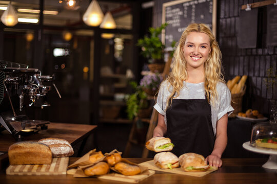 Female barista wearing apron smiling and holding sandwich behind wooden café counter, copy space