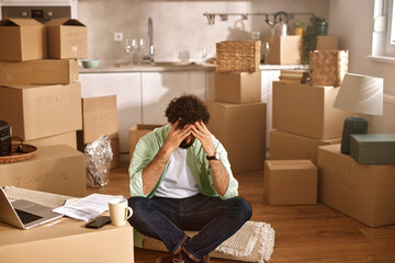 A person sits on the floor among unpacked boxes, displaying frustration while trying to manage the chaos of a recent move. The disorganized kitchen space adds to the feeling of overwhelm.