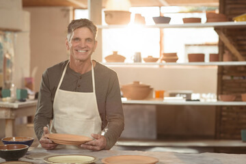 Male ceramic artist smiling while holding clay plate at ceramics workshop wearing canvas apron