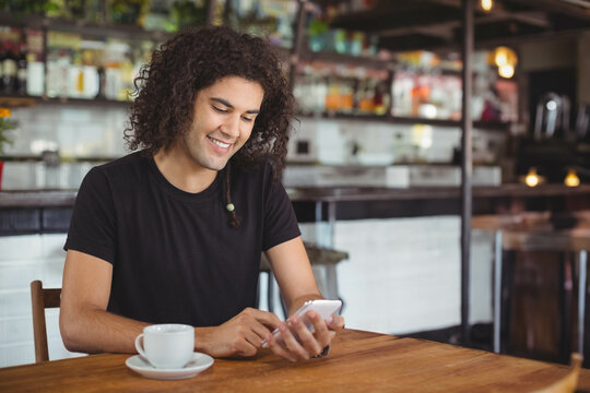 Man sitting at wooden table in cafe smiling at smartphone with ceramic coffee cup and saucer