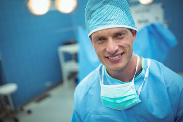 Male surgeon standing in operating room with surgical lights wearing blue scrubs, cap and mask