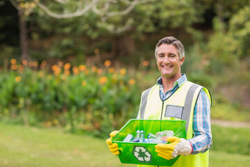 Male recycling worker smiling, holding green bin wearing yellow gloves on park lawn, copy space