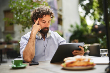 A man with curly hair and headphones focuses intently on a tablet, seated at an outdoor cafe table. A sandwich and coffee are visible in the foreground, indicating a business meeting or work session.