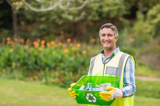 Male recycling worker smiling, holding green bin wearing yellow gloves on park lawn, copy space