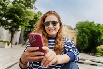 Happy attractive mature mid adult european woman in sunglasses using smartphone mobile phone while walking in summer city