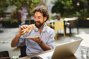 A man with curly hair and headphones smiles while holding a sandwich in one hand and sitting at a table with a laptop outdoors, enjoying his lunch break in a vibrant cafe environment.