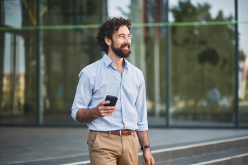 A man dressed in a light blue shirt and beige pants walks with a smile, holding his smartphone,...