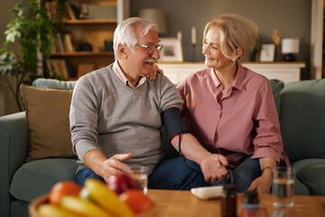 An older man is wearing a blood pressure monitor on his arm while sitting on a couch next to his wife. They are looking at each other and smiling in a cozy living room setting.