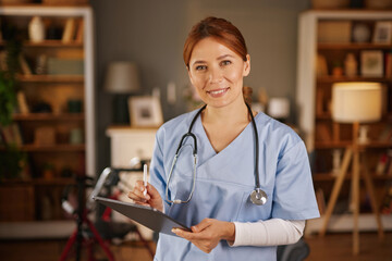 A smiling female healthcare professional, wearing scrubs and stethoscope, stands indoors holding a pen and clipboard, likely documenting patient information during a home visit.