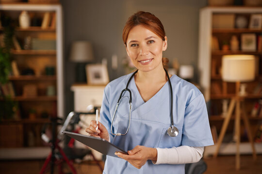 A smiling female healthcare professional, wearing scrubs and stethoscope, stands indoors holding a pen and clipboard, likely documenting patient information during a home visit.