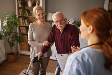 In a living room, a Nurse consults with a senior man using a walker as his wife stands by his side holding his arm. The Nurse is writing notes on a clipboard during the daytime.