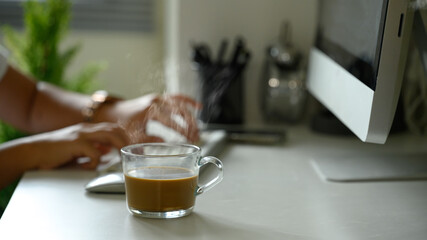 Fresh coffee cup on desk with steam rising, hands woman working in the background.