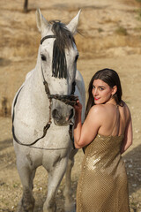 Beautiful young girl in a pretty dress posing with her back to the camera next to a white horse in...