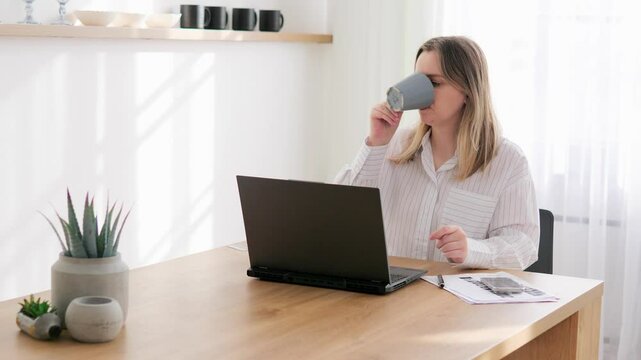 Student is studying online, sitting laptop home. Young woman carefully studies task on screen while taking sip tea. Studying comfortable environment allows student to concentrate master new material.
