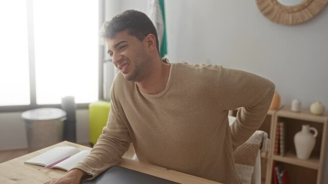 Man back holds posture at home as young hispanic guy sits indoor feeling discomfort and pain.