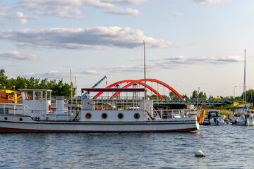Passenger ship moored in a riverside harbor, with a modern red arch bridge and marina with yachts...