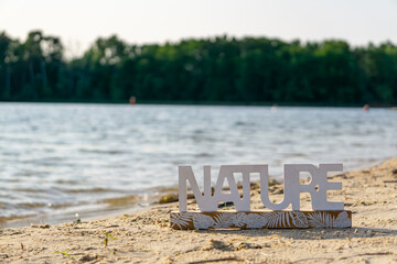 Wooden "NATURE" sign placed on a sandy beach by the lake, with water and a forest line in the background. A calm summer scene emphasizing connection with nature.