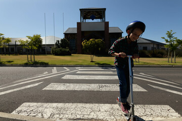 boy riding kick scooter wearing safety helmet across painted crosswalk on suburban road, copy space © WavebreakMediaMicro