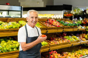Senior male grocery store employee wearing apron holding red tomato beside produce bins