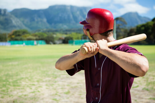 Baseball player holding wooden bat and standing in batter box wearing red helmet on sunlit field