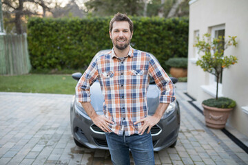 Man in his thirties standing confidently on suburban driveway with gray compact car