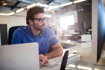 Man in his thirties in blue shirt typing on wireless keyboard at office desk with laptop