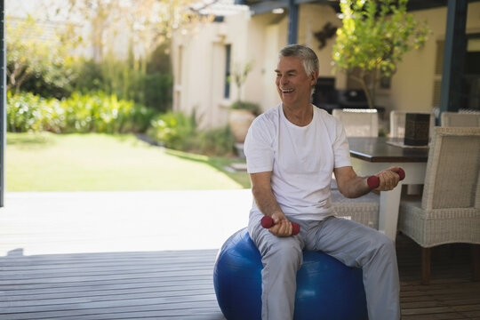 Senior man sitting on blue exercise ball lifting red dumbbells on patio near wicker table, plants