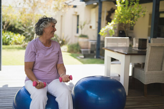 Fototapeta Senior woman balancing on blue exercise ball on wooden patio deck holding pink dumbbells