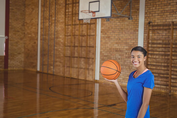 Female teenager posing on court holding orange basketball under hoop with climbing bars, copy space