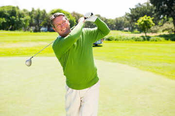 Senior male golfer swinging driver above shoulder on putting green with golf cart and glove