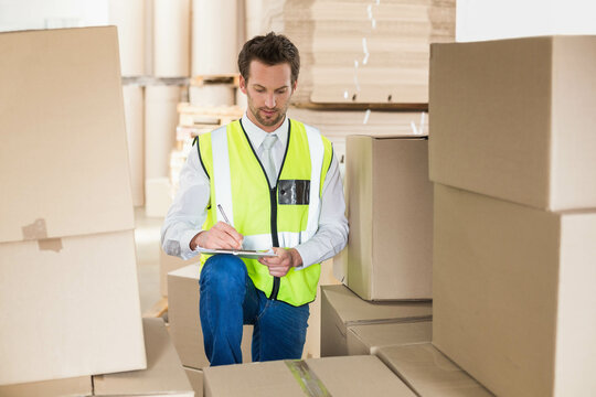 Man kneeling in warehouse writing stock count with pen on clipboard among cardboard boxes, pallets