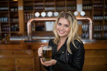 Woman in her twenties holding beer mug smiling toward camera at metal beer taps in bar