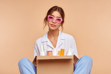 Woman holding a box of various skincare products. She is wearing pink sunglasses and pajamas while sitting against a beige background.