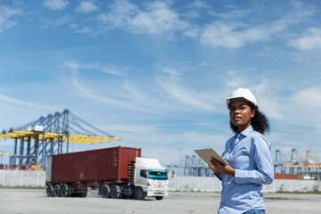 A female logistics officer in a hard hat observes cargo operations at a shipping port. Holding a tablet, she monitors container trucks and cranes involved in global transport and export logistics.