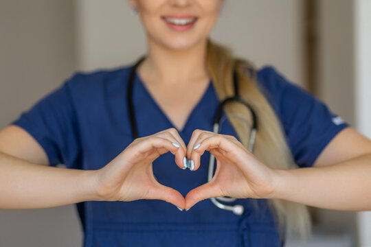 A healthcare professional in a blue uniform smiles warmly and gestures a heart symbol, demonstrating love, empathy, and dedication to patient care