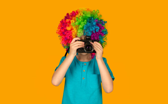 Boy using a cameras. Funny boy in multi-coloured wig. Smiling little boy in clown wig. Little boy making photo on yellow background. Little boy on a taking a photo using a vintage camera