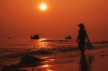 Silhouette of a Vietnamese woman in a straw hat on shore of sea with ships in a fishing village in morning at sunrise in Vietnam