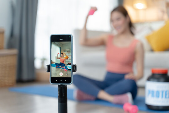 Asian fitness influencer presenting whey protein during online workout at home.Asian female influencer recording online fitness video at home,introducing whey protein supplement.Concept of health care - Powered by Adobe