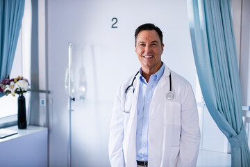 Doctor wearing stethoscope standing in hospital bay 2 beside IV pole and flowers near blue curtains