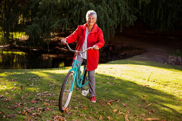 Senior woman holding teal bicycle on sunlit lawn with autumn leaves near pond under evergreen trees